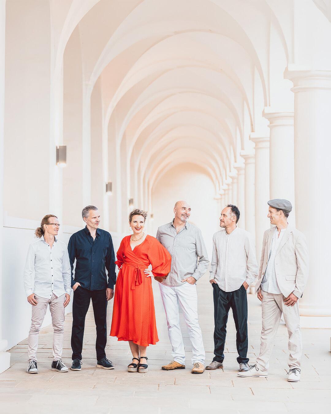 tumba-ito, sechsköpfige band mit sängerin im roten kleid vor heller säulenkulisse