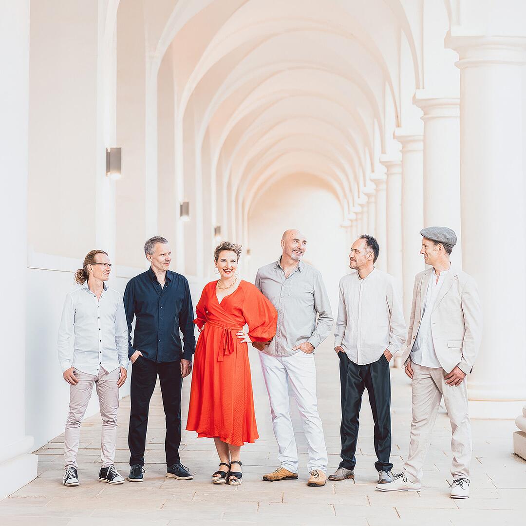 tumba-ito, sechsköpfige band mit sängerin im roten kleid vor heller säulenkulisse