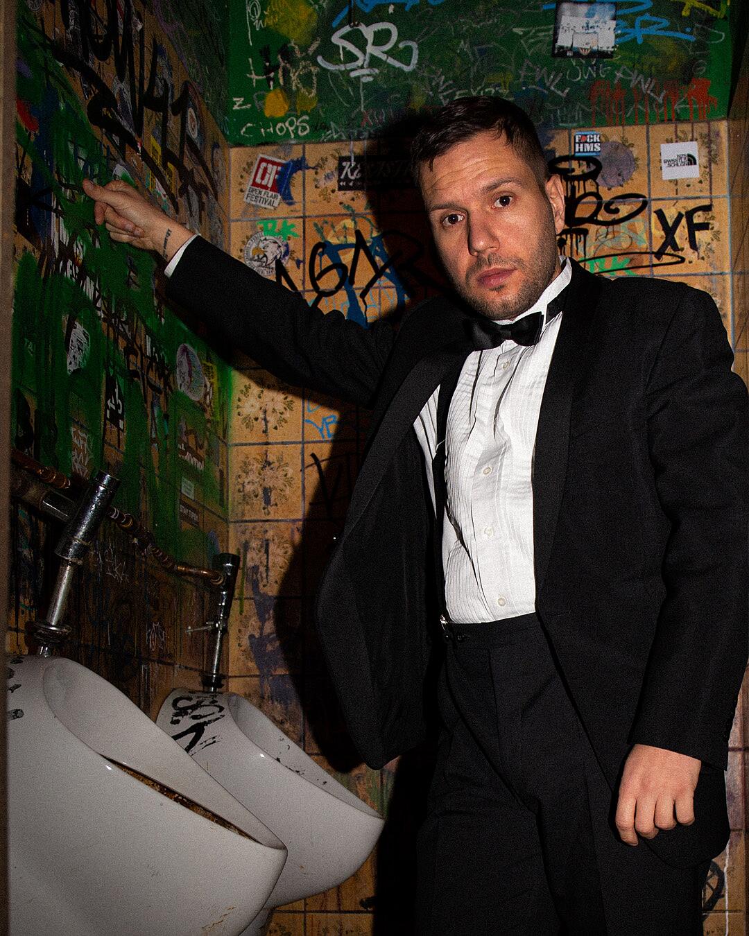 MC Bomber, Mann im Anzug vor Graffitiwand in Toilette