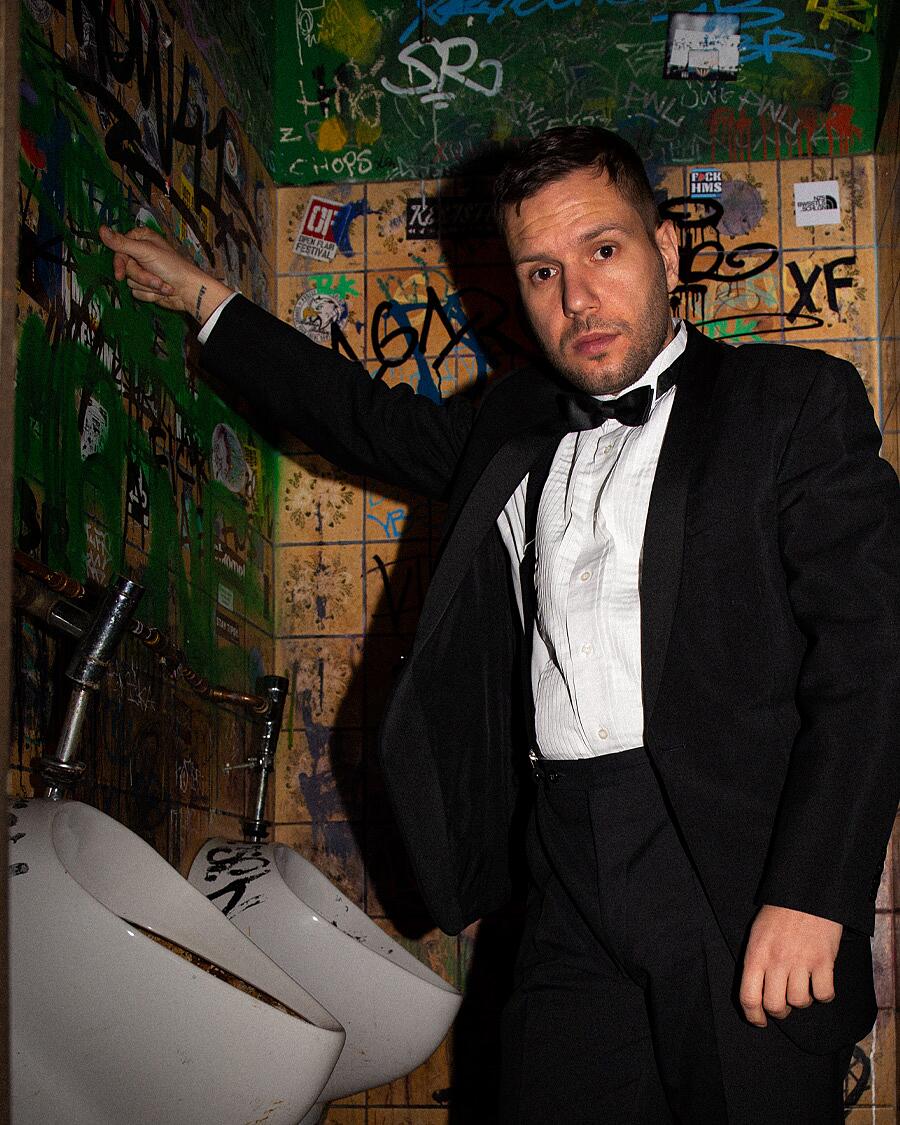 MC Bomber, Mann im Anzug vor Graffitiwand in Toilette