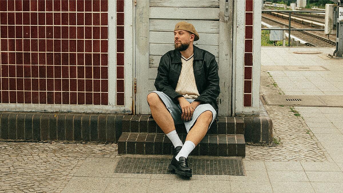 Luca Noel, Mann sitzt vor Gebäude an Bahnsteig